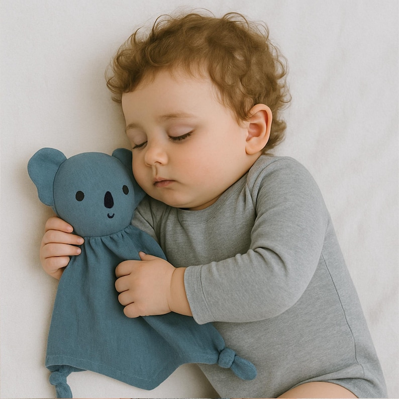Pu&ograve; includere: Un bambino che dorme abbraccia un tenero koala blu. Il giocattolo morbido ha un volto sorridente e angoli annodati. Il bambino indossa una tutina grigia a maniche lunghe e riposa su una superficie bianca.