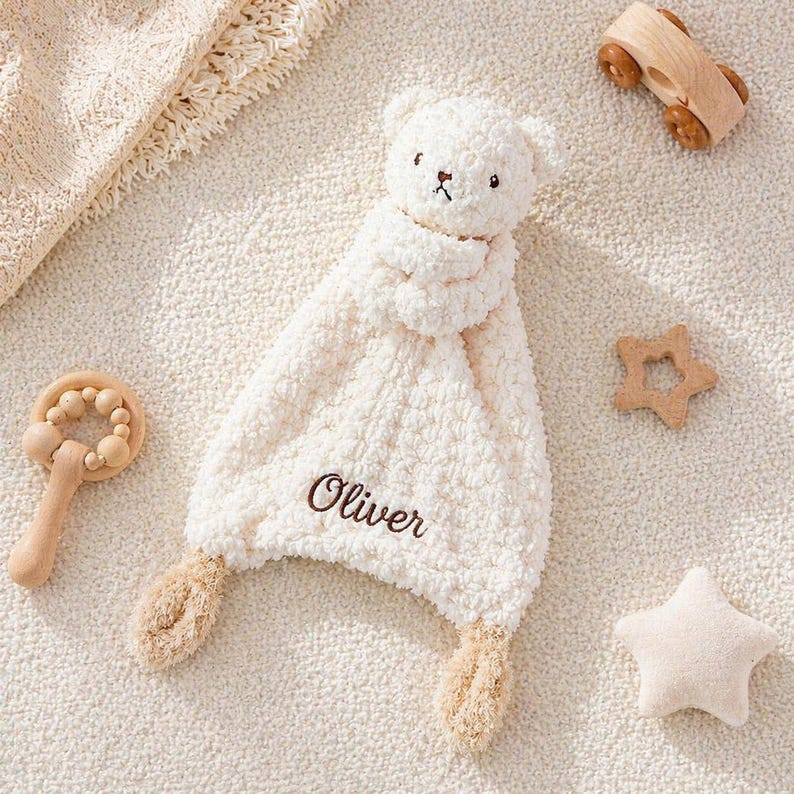 May include: A cream-colored baby blanket with a teddy bear head and the name "Oliver" embroidered in brown. The blanket has two tan-colored ear-shaped extensions. Wooden toys, including a car, rattle, and star-shaped objects, are scattered around the blanket.
