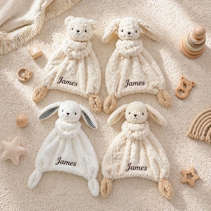 May include: Four plush baby security blankets in cream and white, each featuring a stuffed animal head and the name "James" embroidered on the blanket. Two are bear-shaped, and two are bunny-shaped. Additional wooden toys are scattered around the blankets.