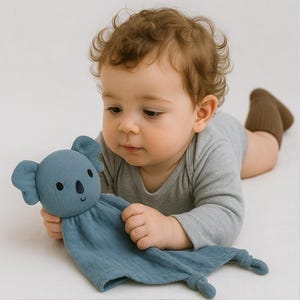 Pu&ograve; includere: Un bambino tiene in mano una coperta di sicurezza a forma di koala blu. Il koala ha occhi neri e un piccolo sorriso. Il bambino indossa un body grigio e calzini marroni. Lo sfondo &egrave; bianco.