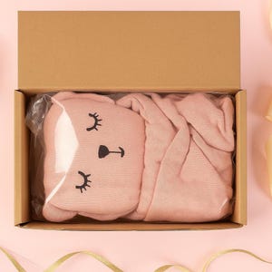 Pu&ograve; includere: Un set di coperta e cuscino rosa per bambini in una scatola di cartone. Il cuscino ha la forma di una faccia di coniglietto con ciglia e naso neri. La coperta &egrave; piegata ordinatamente accanto al cuscino. La scatola &egrave; aperta, rivelando il contenuto.