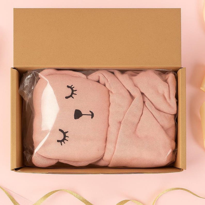 Pu&ograve; includere: Un set di coperta e cuscino rosa per bambini in una scatola di cartone. Il cuscino ha la forma di una faccia di coniglietto con ciglia e naso neri. La coperta &egrave; piegata ordinatamente accanto al cuscino. La scatola &egrave; aperta, rivelando il contenuto.