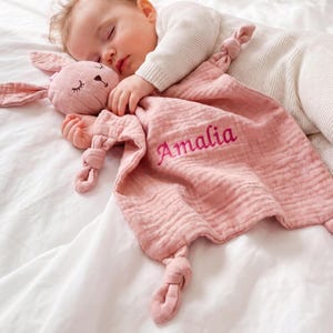 Pu&ograve; includere: Un bambino che dorme riposa con una coperta di sicurezza a forma di coniglietto rosa. La coperta &egrave; personalizzata con il nome "Amalia" ricamato in filo rosa abbinato. Il coniglietto ha occhi ricamati e un'espressione dolce.