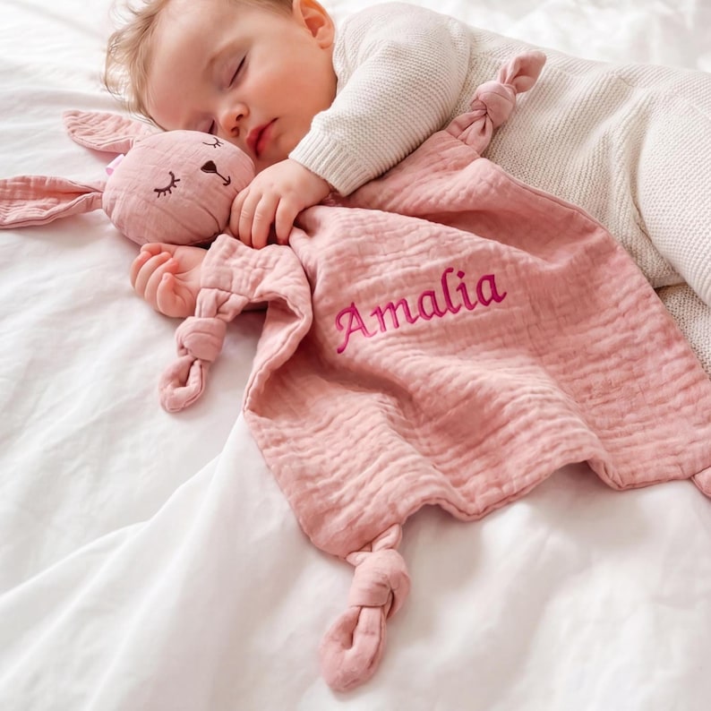 Pu&ograve; includere: Un bambino che dorme riposa con una coperta di sicurezza a forma di coniglietto rosa. La coperta &egrave; personalizzata con il nome "Amalia" ricamato in filo rosa abbinato. Il coniglietto ha occhi ricamati e un'espressione dolce.