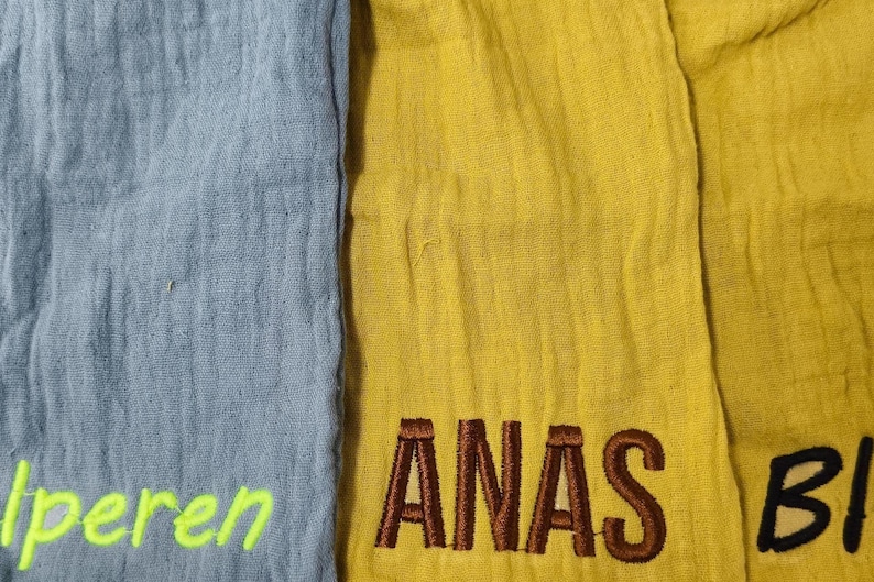 Pu&ograve; includere: Primo piano di tre sezioni di tessuto in blu, giallo e giallo-verde. La sezione gialla ha la parola ricamata "ANAS" in marrone. La sezione blu ha la parola ricamata "Iperen" in verde neon. La sezione giallo-verde ha la parola ricamata "BI" in nero.