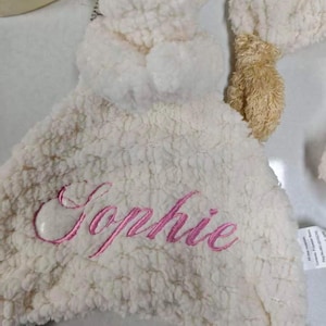May include: A white, plush baby blanket with the name "Sophie" embroidered in pink script. The blanket has a soft, textured surface and a small, tan teddy bear head attached.