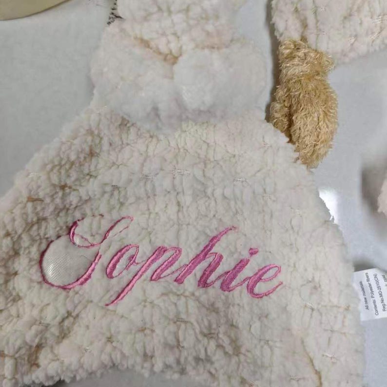 May include: A white, plush baby blanket with the name "Sophie" embroidered in pink script. The blanket has a soft, textured surface and a small, tan teddy bear head attached.