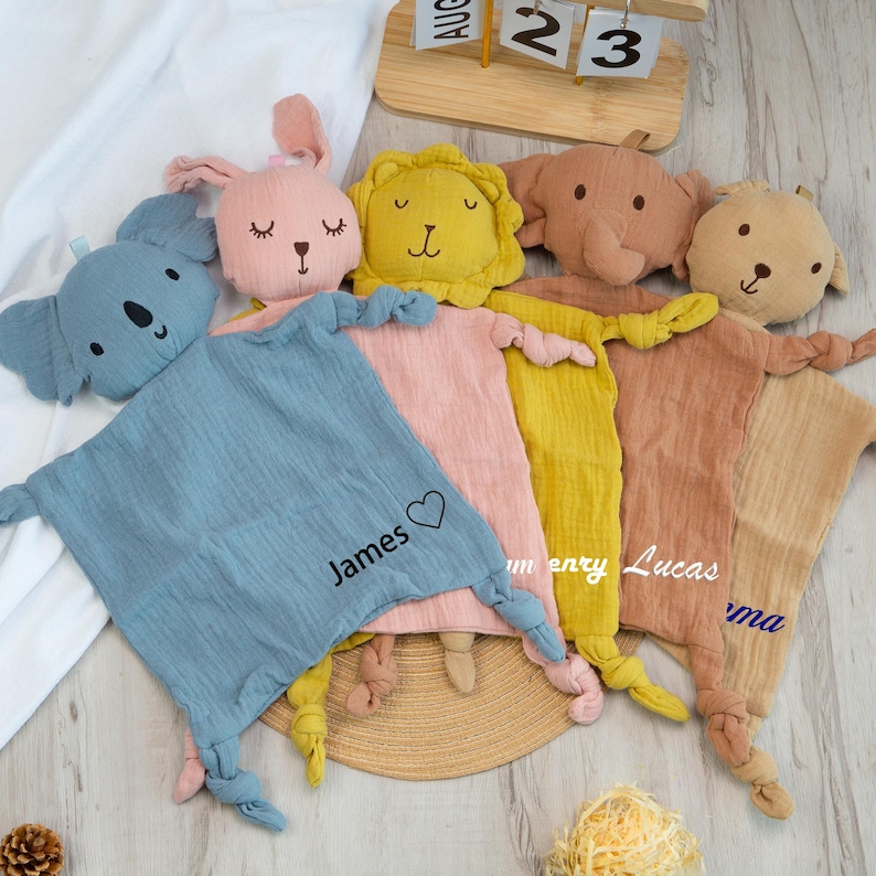 Pu&ograve; includere: Una collezione di morbide e soffici copertine di sicurezza per bambini in vari colori, ognuna con una testa di animale imbottita. Le coperte presentano teste di animali tra cui un koala, un coniglio, un leone, un elefante e un orso. Alcune coperte hanno nomi.