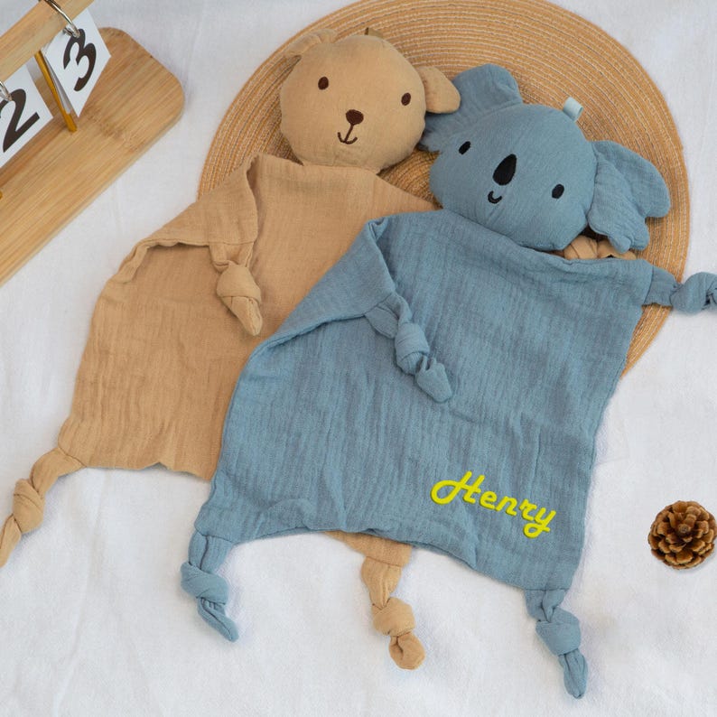 Pu&ograve; includere: Due doudou per bambini, uno beige e uno blu, ciascuno con una testa di animale di peluche. Il doudou beige ha una testa di orso, e il doudou blu ha una testa di koala. Il doudou blu ha il nome "Henry" ricamato in giallo.