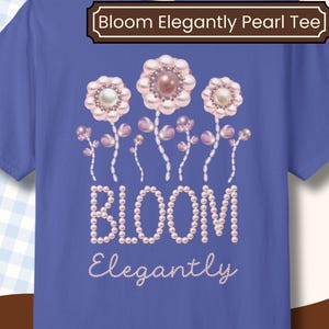 May include: A periwinkle blue t-shirt with a floral design made of pearls and the words "BLOOM Elegantly" in a script font. The top of the shirt has the text "Bloom Elegantly Pearl Tee" in a brown banner.
