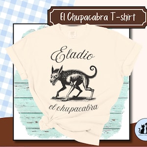 Puede incluir: Camiseta color crema con una ilustración negra de un Chupacabra, con el texto "Eladio el chupacabra" en un estilo vintage. La camiseta tiene cuello redondo clásico y mangas cortas, ideal para uso casual.