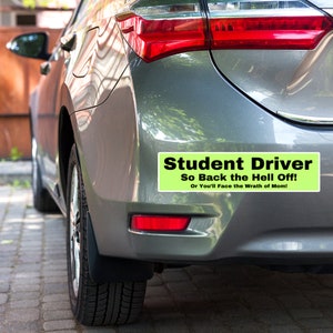 May include: A green and black bumper sticker that reads "Student Driver So Back the Hell Off! Or You'll Face the Wrath of Mom!" on a silver car.
