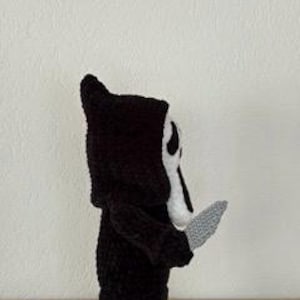 Pu&ograve; includere: Figura all'uncinetto di un personaggio con una veste nera con cappuccio e una maschera bianca con fori neri per occhi e bocca. La figura tiene in mano un coltello grigio. La figura &egrave; su una superficie chiara su sfondo bianco.