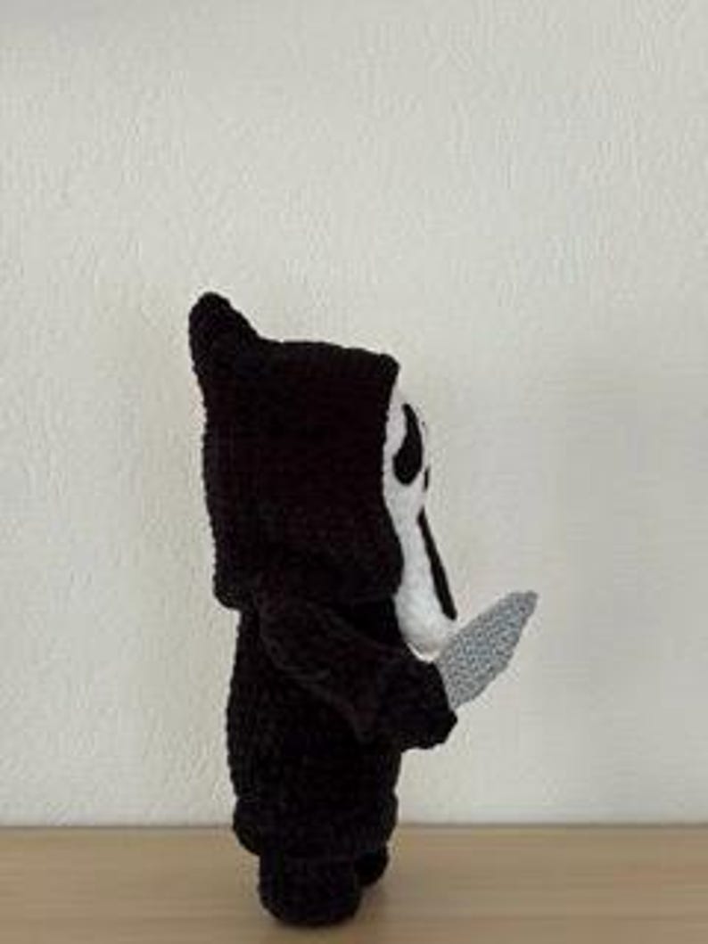 Pu&ograve; includere: Figura all'uncinetto di un personaggio con una veste nera con cappuccio e una maschera bianca con fori neri per occhi e bocca. La figura tiene in mano un coltello grigio. La figura &egrave; su una superficie chiara su sfondo bianco.