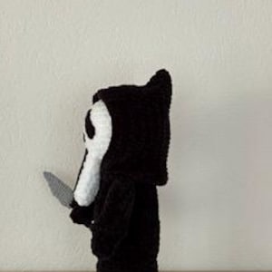Pu&ograve; includere: Bambola all'uncinetto in bianco e nero fatta a mano a forma di personaggio del film Scream, con un coltello d'argento. La bambola ha una maschera bianca con fori neri per gli occhi e una veste con cappuccio nera. La bambola &egrave; su una superficie di legno.