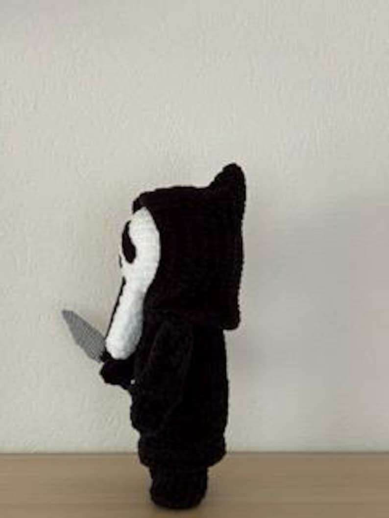 Pu&ograve; includere: Bambola all'uncinetto in bianco e nero fatta a mano a forma di personaggio del film Scream, con un coltello d'argento. La bambola ha una maschera bianca con fori neri per gli occhi e una veste con cappuccio nera. La bambola &egrave; su una superficie di legno.
