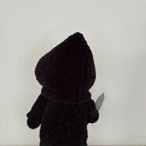 Pu&ograve; includere: Una figura nera all'uncinetto di un personaggio incappucciato che tiene in mano un coltello d'argento. La figura &egrave; vista da dietro, evidenziando la consistenza del filato e la forma del cappuccio e del mantello. Lo sfondo &egrave; un muro bianco.