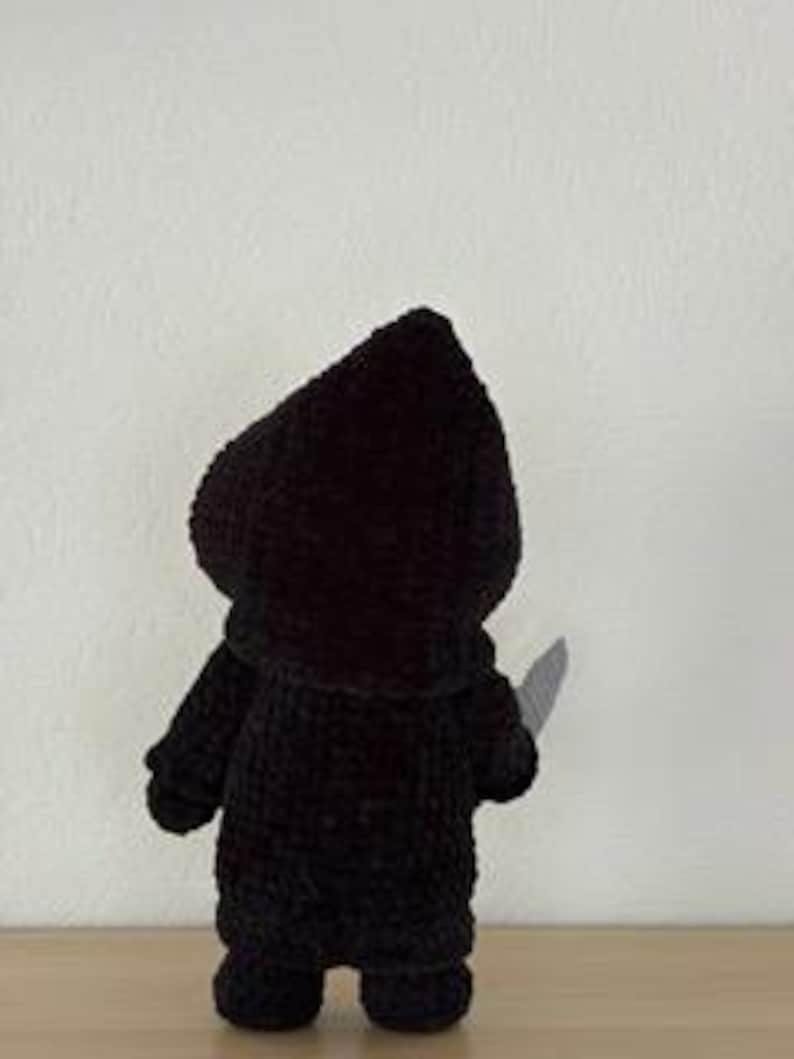 Pu&ograve; includere: Una figura nera all'uncinetto di un personaggio incappucciato che tiene in mano un coltello d'argento. La figura &egrave; vista da dietro, evidenziando la consistenza del filato e la forma del cappuccio e del mantello. Lo sfondo &egrave; un muro bianco.