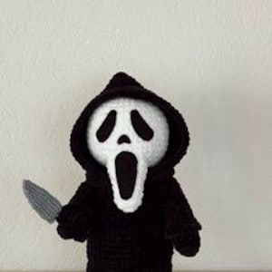Pu&ograve; includere: Bambola all'uncinetto fatta a mano del personaggio di Ghostface della serie di film horror Scream. La bambola indossa un abito con cappuccio nero e tiene in mano un coltello grigio. Il viso della bambola &egrave; bianco con dettagli neri per occhi e bocca.