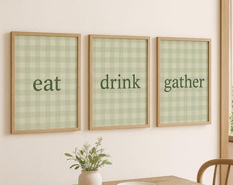 May include: Three framed wall art pieces with the words "eat", "drink", and "gather" in dark green lettering. Each piece has a light green and white gingham pattern background and a natural wood frame. The art is displayed above a wooden table.