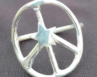 May include: A silver-colored ring featuring a peace symbol design with a star in the center. The ring has a circular shape with three lines extending from the center, and a star in the middle. The ring is set against a dark background.