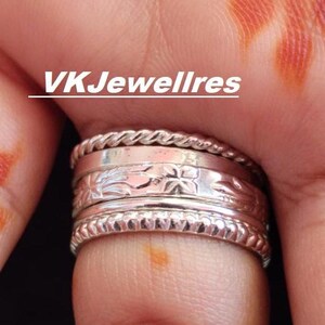 Può includere: Un anello d'argento su un dito, con più fasce e disegni intricati. L'anello presenta un motivo floreale e una texture a corda intrecciata. Il testo "VKJewellres" è visualizzato sopra l'anello.