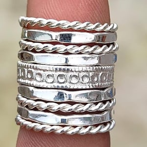May include: A collection of silver rings stacked on a finger. The rings have various designs, including twisted rope, smooth bands, and a band with a repeating pattern of circles and dots. The rings are displayed in a close-up shot.