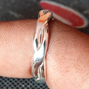 May include: A close-up of a silver ring with a braided design. The ring is smooth and shiny, reflecting light. The ring is on a finger.