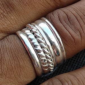 May include: A collection of silver rings stacked on a finger. The rings include smooth bands and a braided rope design. The rings are made of a shiny, reflective metal.