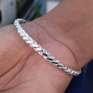 Könnte beinhalten: Ein silberfarbenes Armband mit geflochtenem Design. Das Armband wird an einem Handgelenk getragen, wodurch sein kompliziertes Muster und seine reflektierende Oberfläche hervorgehoben werden. Das Schmuckstück scheint aus Metall zu sein, ein stilvolles Accessoire.