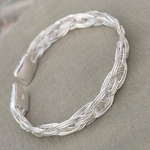 May include: A silver-coloured braided cuff bracelet. The bracelet features a woven design with a clasp closure. The metal has a bright, reflective finish, and the bracelet is open at one end.