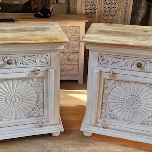 Puede incluir: Dos mesitas de noche de madera blanca con diseños florales tallados. Cada mesita tiene una tapa de madera de color claro, un cajón con un pomo de latón y un gran diseño de sol tallado en la puerta. Las mesitas tienen patas cortas y redondeadas.
