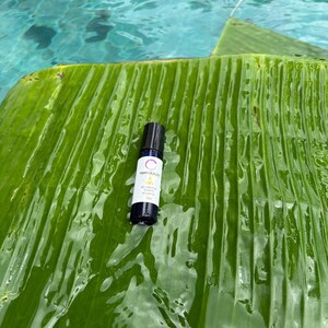 May include: A small, white and black essential oil roller bottle with the word "Tranquility" printed on it. The bottle is on a large, green banana leaf, with a blue water background.