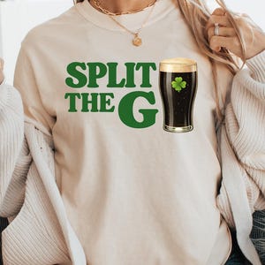 May include: Beige t-shirt with the text "SPLIT THE G" in green, and a graphic of a dark stout beer with a shamrock. The person is holding a glass of a light brown beverage. The shirt is a casual style.