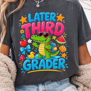 Könnte beinhalten: Dunkelgraues T-Shirt mit dem Text "Later Third Grader" und einem Cartoon-Alligator mit Sonnenbrille und Rucksack. Das Design umfasst eine Wassermelonenscheibe, einen Apfel und eine Quietscheente. Lässiger Stil.
