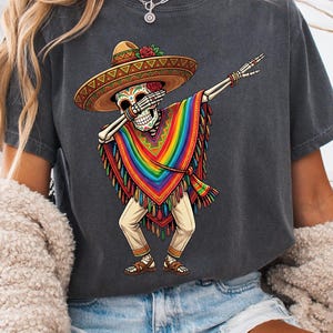 Könnte beinhalten: Dunkelgraues T-Shirt mit einem Skelett, das einen Sombrero und einen Regenbogen-Poncho trägt und dabbt. Das Skelett hat ein Totenkopfgesicht und einen Hut mit einer roten Rose. Der Poncho hat ein Regenbogenmuster. Das T-Shirt ist lässig.