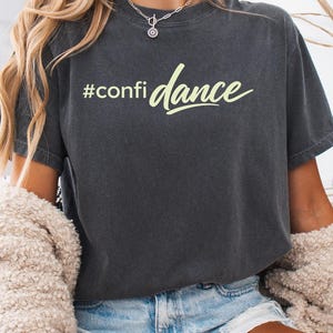 May include: Dark gray t-shirt with the text "#confidance" in a light green script. The shirt is paired with denim shorts and a cream-colored sweater. The overall aesthetic is casual and comfortable.