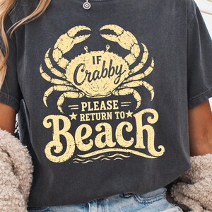 以下が含まれることがあります： IF CRABBY PLEASE RETURN TO BEACHというテキストとカニの黄色いグラフィックプリントが施された、チャコールグレーの半袖Tシャツ。クルーネックでゆったりとしたフィット感があり、ライトブルーのデニムショートパンツと組み合わされています。
