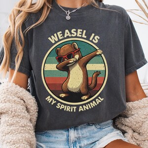 Puede incluir: Camiseta gris oscuro con un dibujo de una comadreja de dibujos animados con gafas de sol y haciendo un dab, con el texto "WEASEL IS MY SPIRIT ANIMAL" en un diseño circular. La comadreja es marrón con vientre beige.