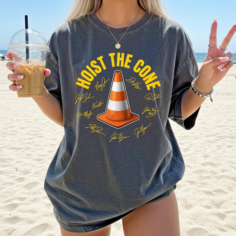 May include: Dark gray t-shirt with the text "HOIST THE CONE" in yellow, surrounding an orange and white traffic cone graphic. The shirt features multiple signatures in yellow. The shirt is a relaxed fit.