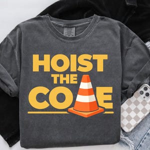 May include: Dark gray sweatshirt with the words "HOIST THE CONE" in yellow block letters. An orange and white traffic cone graphic is centered below the text. The sweatshirt is on a light-colored surface.