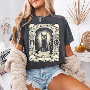 May include: Dark gray t-shirt featuring a black cat illustration within a decorative frame with skulls, a crescent moon, and a star. The cat sits atop a pedestal with a banner below. The design has a vintage, gothic aesthetic.