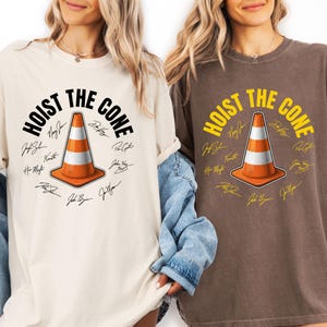 May include: Two t-shirts, one cream and one brown, featuring the text "HOIST THE CONE" above an orange and white traffic cone graphic. The design includes signatures around the cone. The shirts are casual wear.
