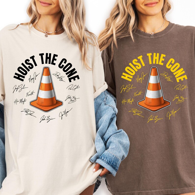 May include: Two t-shirts, one cream and one brown, featuring the text "HOIST THE CONE" above an orange and white traffic cone graphic. The design includes signatures around the cone. The shirts are casual wear.