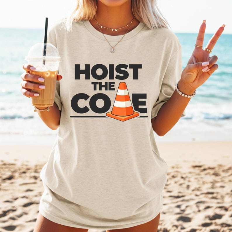May include: Beige t-shirt with the text "HOIST THE CONE" in black letters and a graphic of an orange and white traffic cone. The shirt is worn on a beach with the ocean in the background.