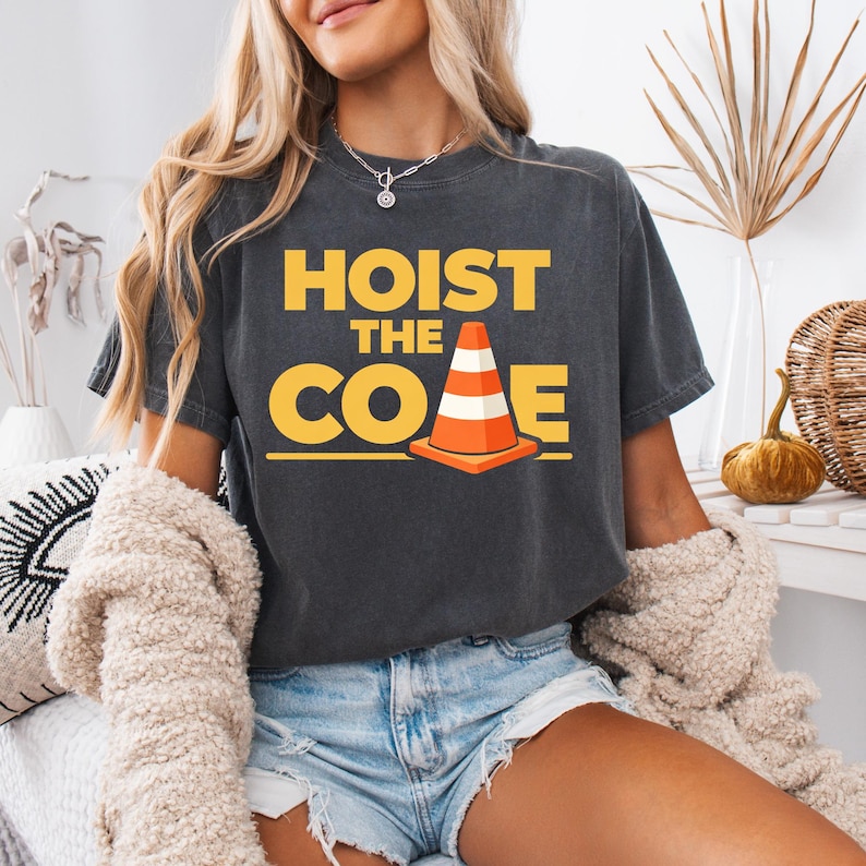 May include: Dark gray t-shirt with the words "HOIST THE CONE" in yellow block letters and a graphic of an orange and white traffic cone. The shirt is a casual style.