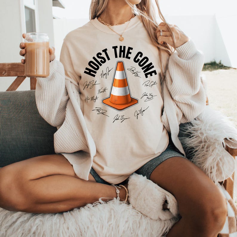 May include: Cream-colored sweatshirt with the text "HOIST THE CONE" in a semi-circle above a traffic cone graphic. The cone is orange and white, with several signatures around it. The sweatshirt is a relaxed fit.