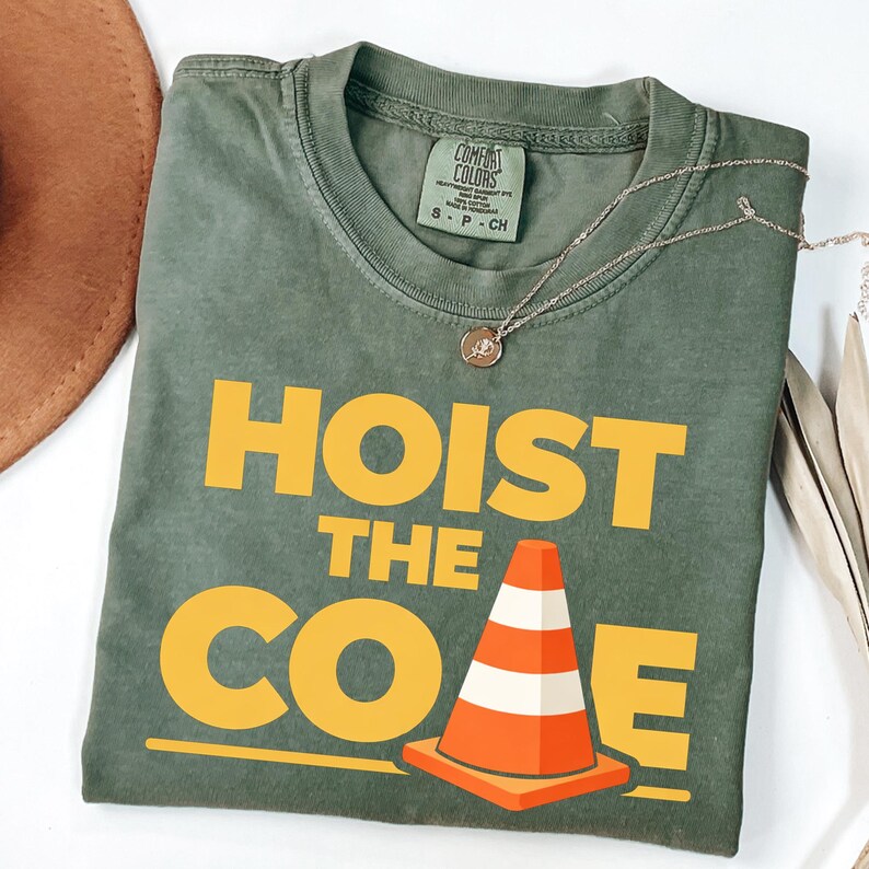 May include: An olive green t-shirt with the words "HOIST THE CONE" in yellow block letters and a graphic of an orange and white traffic cone. A silver necklace with a small pendant is draped across the shirt.