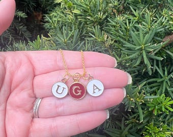 May include: Gold chain necklace with three charms, each featuring a white enamel circle with a gold letter inside. The letters spell "UGA" and the middle charm is red.