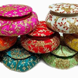May include: A selection of vibrant, round fabric boxes with lids. The boxes are in a range of colours, such as pink, orange, red, white, yellow, teal, green, and navy. Each box features gold embroidery, giving them a decorative and ornate look.